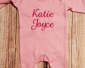 May include: Pink long-sleeved baby onesie with the name "Katie Joyce" embroidered in pink on the front.