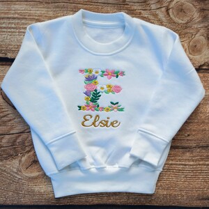 May include: White sweatshirt with a floral embroidered letter E and the name Elsie in gold embroidery.
