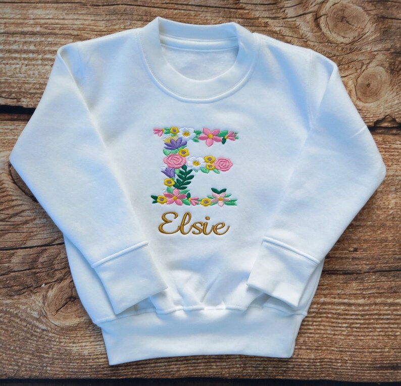 May include: White sweatshirt with a floral embroidered letter E and the name Elsie in gold embroidery.