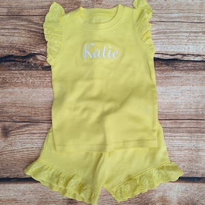 May include: Yellow sleeveless top and shorts set with white lace trim and the name "Katie" embroidered on the front of the top.