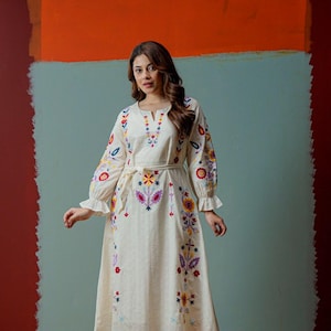 Puede incluir: Vestido bordado color crema con mangas largas y cinturón anudado. El vestido presenta bordados florales coloridos en las mangas, el corpiño y la falda. El fondo es una pared de dos tonos con secciones naranja y verde azulado.