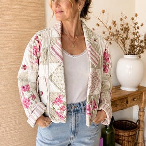May include: A quilted jacket featuring a patchwork design with floral patterns in shades of pink, cream, and beige. The jacket has long sleeves with folded cuffs, a shawl collar, and a front opening, worn over a white tank top and light blue jeans.