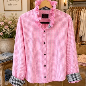 May include: A long-sleeved button-down shirt in a pink and white gingham pattern. The shirt features a ruffled collar, black buttons, and contrasting black and white checkered fabric on the inside of the collar and the wide, folded-up cuffs.