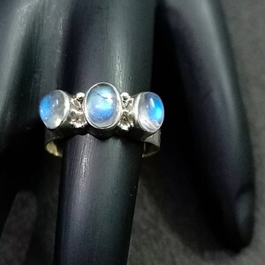 May include: A silver ring with three oval-shaped moonstone gemstones. The stones are set in a silver band and have a light blue iridescent sheen.