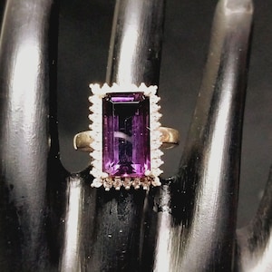 May include: A gold ring with a large, rectangular, purple amethyst gemstone surrounded by smaller diamonds.