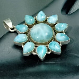 May include: A silver pendant with a blue and white gemstone in the center surrounded by smaller blue and white gemstones. The pendant is shaped like a flower.