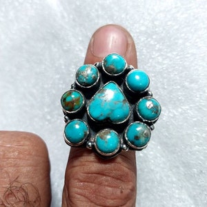 Op de afbeelding: Een zilveren ring met een cluster van turquoise stenen. De stenen zijn in een hartvorm gezet en worden omringd door kleinere stenen.