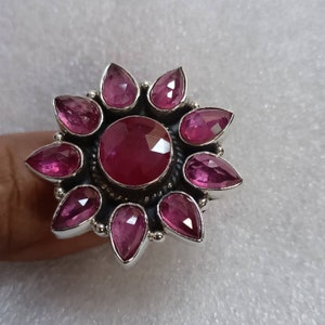 May include: A silver ring with a large pink gemstone in the center surrounded by eight smaller pink gemstones. The ring is shaped like a flower.