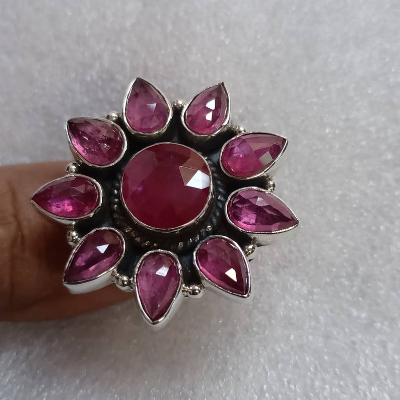 May include: A silver ring with a large pink gemstone in the center surrounded by eight smaller pink gemstones. The ring is shaped like a flower.
