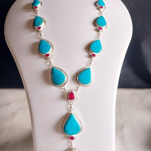 May include: A turquoise and silver necklace with teardrop-shaped turquoise stones and red accents. The necklace is displayed on a white mannequin.