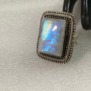 Może przedstawiać: Srebrny pierścionek z dużym, prostokątnym, opalizującym kamieniem księżycowym osadzonym w oprawie z koralików.