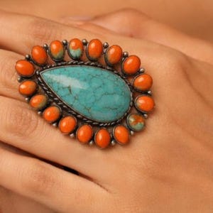 Może przedstawiać: Duży, owalny pierścionek z turkusowym kamieniem pośrodku, otoczony pomarańczowymi kamieniami. Pierścionek ma srebrną oprawę. Wyrazista biżuteria.