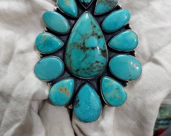 May include: A turquoise pendant with a large central stone surrounded by smaller stones in a teardrop shape.