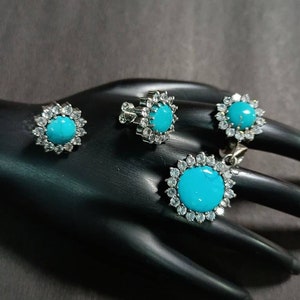 May include: A turquoise gemstone jewelry set with silver settings and clear cubic zirconia stones. The set includes a pendant, a pair of earrings, and a ring.