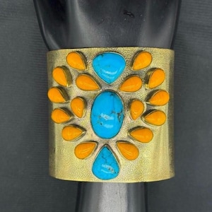 May include: A wide gold-toned metal cuff bracelet featuring a central oval turquoise stone, a smaller teardrop turquoise stone at the bottom, and a top teardrop turquoise stone, all surrounded by numerous small, bright orange teardrop-shaped stones.