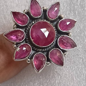Op de afbeelding: Een zilveren ring met een grote roze edelsteen omringd door kleinere roze edelstenen in de vorm van een bloem.