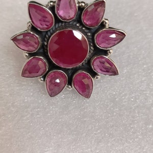 May include: A silver ring with a large, round, red gemstone surrounded by eight smaller, teardrop-shaped, pink gemstones.