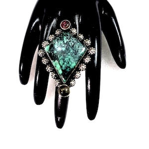 Może przedstawiać: Srebrny pierścionek z dużym, zielonym turkusem osadzonym w kwadratowym kształcie. Pierścionek jest ozdobiony mniejszymi kamieniami i misternymi detalami.