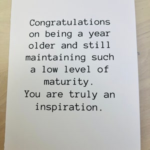 May include: A white card with black text that reads "Congratulations on being a year older and still maintaining such a low level of maturity. You are truly an inspiration."