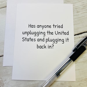 May include: A white greeting card with the text "Has anyone tried unplugging the United States and plugging it back in?" is next to a white envelope and a black and silver pen on a light wood surface.