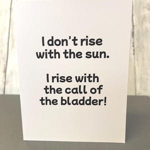 May include: A white greeting card with black text. The text reads: "I don't rise with the sun. I rise with the call of the bladder!" The card is on a gray surface.