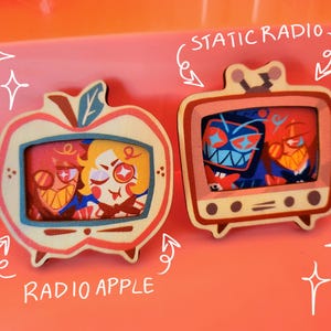Può includere: Due spille di legno a forma di televisore e mela. La spilla del televisore ha un personaggio dei cartoni animati rosso e blu all'interno con il testo "Static Radio". La spilla della mela ha due personaggi dei cartoni animati all'interno con il testo "Radio Apple".