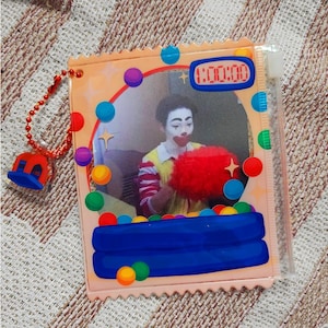 Pode incluir: Um porta-fotos de plástico transparente com uma imagem de palhaço, um design colorido de piscina de bolinhas e um pingente vermelho e azul. A foto mostra uma pessoa vestida de palhaço. O suporte tem um visor de relógio digital mostrando 1:00:00.