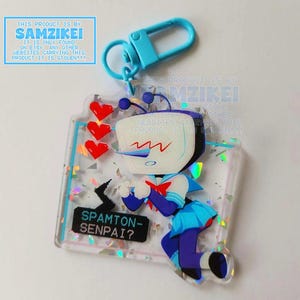 Pode incluir: Um porta-chaves de acrílico transparente com uma personagem com cabeça de televisão, vestida com um traje de marinheiro azul e branco. O design inclui três corações vermelhos pixelizados, um balão de fala preto com o texto SPAMTON-SENPAI? e detalhes de glitter holográfico. Está preso a um mosquetão de plástico azul claro.