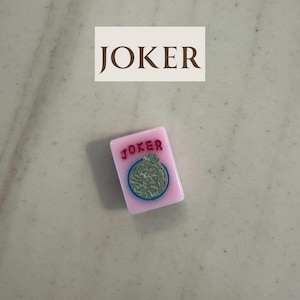 May include: A pink Mahjong tile with the word "JOKER" in red at the top. The tile features a green and blue circular design. Above the tile is a white rectangle with the word "JOKER" in brown.