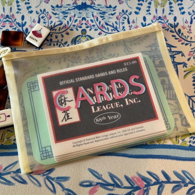 May include: A clear zippered pouch containing a deck of cards with the text "CARDS LEAGUE, INC." and "OFFICIAL STANDARD HANDS AND RULES". The pouch also holds a light green card with a geometric design. A few Mahjong tiles are visible.
