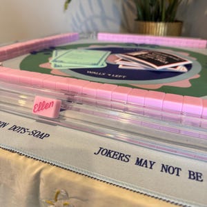 May include: A pink Mahjong set with tiles, racks, and game cards. The game board has a green and blue design. One tile is labeled "Ellen" in pink script. The game is set up on a light blue cloth.