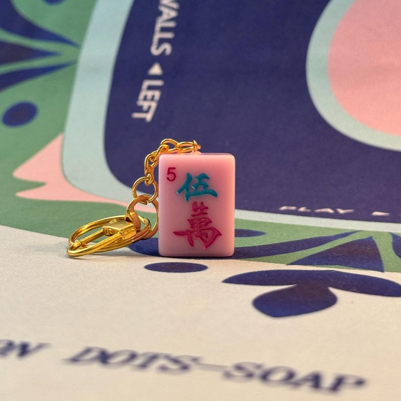May include: A pink Mahjong tile keychain with gold-colored hardware. The tile features the number 5 and Chinese characters in green and pink. The background is a blue and green patterned surface.