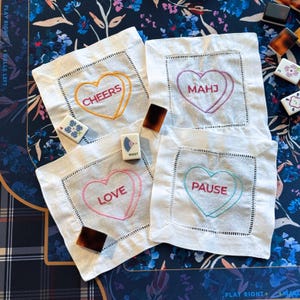 May include: Four white linen coasters with embroidered heart designs. Each coaster features a different word: "CHEERS" in orange, "MAHJ" in purple, "LOVE" in pink, and "PAUSE" in teal. The coasters are square with a stitched border.