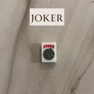 May include: A white Mahjong tile with the word "JOKER" in red and a green circular design. Above the tile is a rectangular sign with the word "JOKER" in brown.