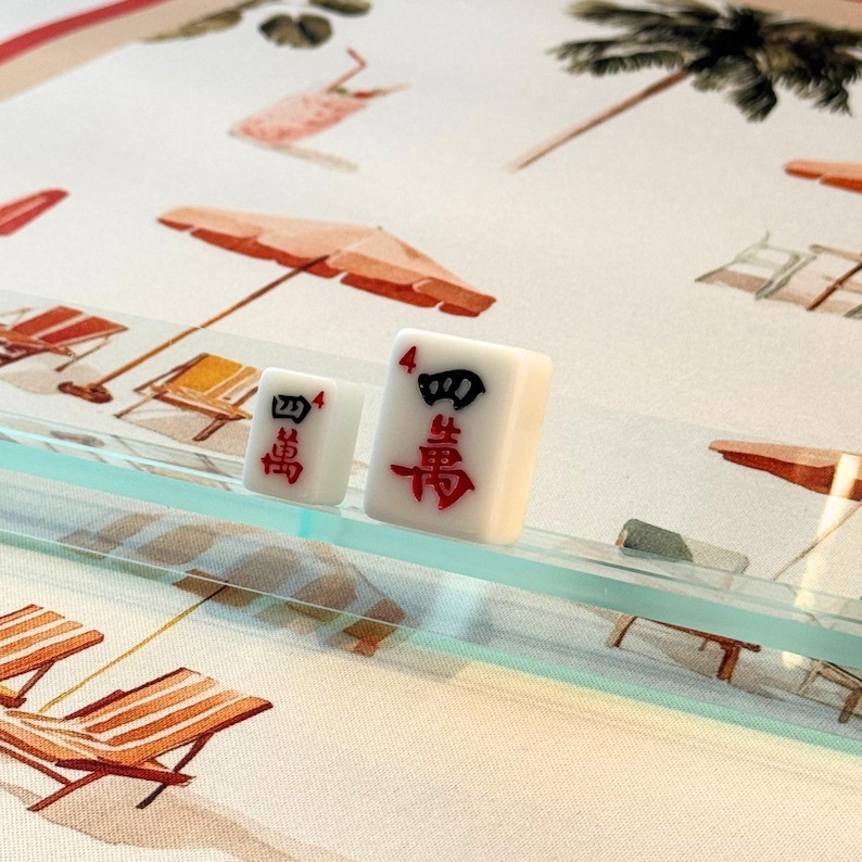 May include: Two white Mahjong tiles with red and black characters, displaying the number four. The tiles are on a clear glass surface, with a beach-themed background featuring umbrellas and lounge chairs.