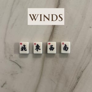 May include: Four white Mahjong tiles with black Chinese characters and red accents, labeled N, E, W, and S, representing the cardinal directions. The word "WINDS" is above the tiles. The tiles are on a marble surface.