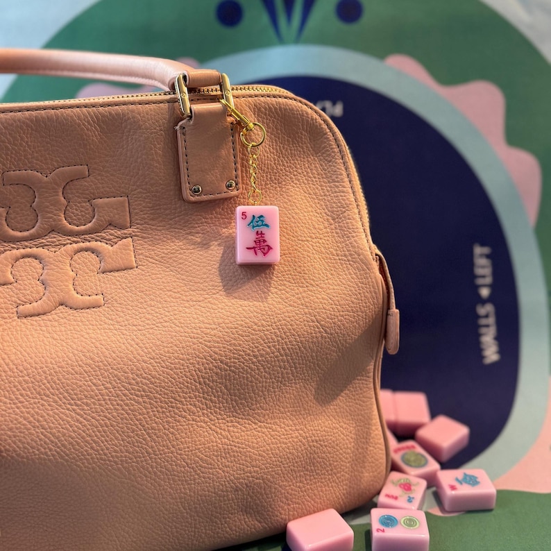 May include: A light pink leather handbag with a gold-tone clasp and a small pink Mahjong tile charm. The charm is attached with a gold chain and features Chinese characters and the number 5. Several loose pink Mahjong tiles are scattered nearby.