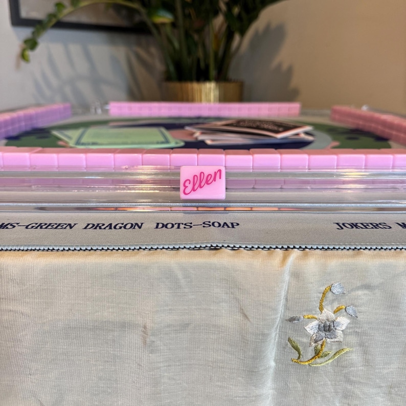 May include: A pink Mahjong set with tiles and racks, featuring the name "Ellen" on a tile. The set is arranged on a table with a floral embroidered cloth. The text "MS-GREEN DRAGON DOTS-SOAP" is visible.