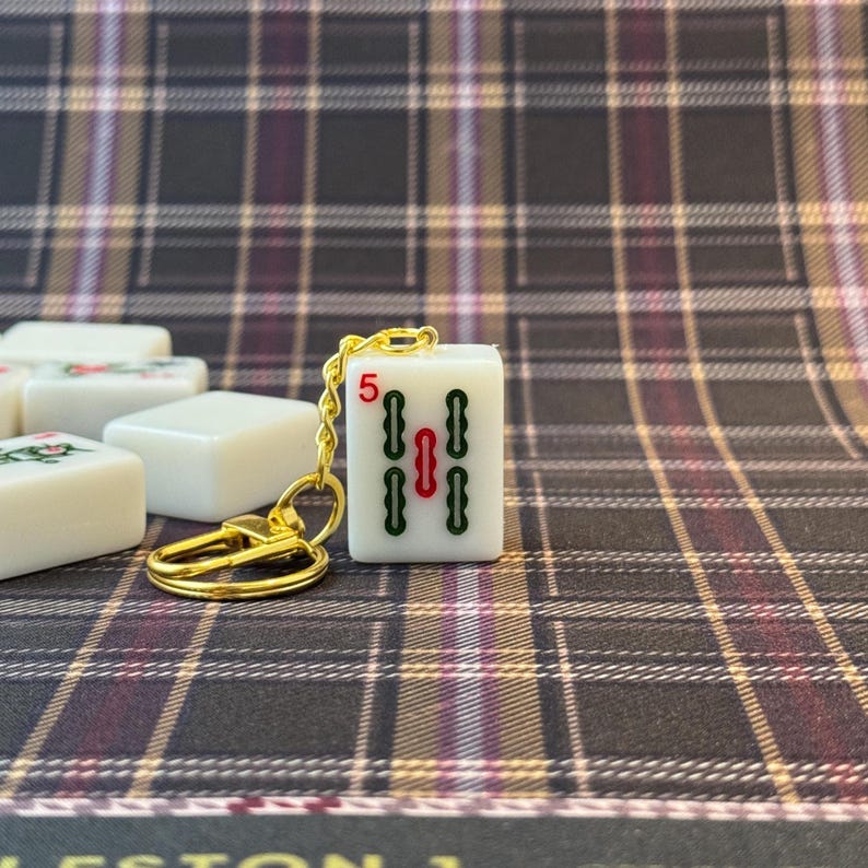 May include: A white Mahjong tile keychain with a gold-colored keyring. The tile displays the number 5 in red and green symbols. Other Mahjong tiles are in the background. The background is a plaid pattern in shades of brown, burgundy, and cream.