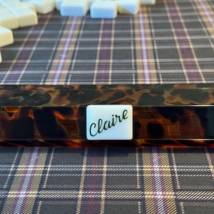 Puede incluir: Un soporte rectangular para fichas de juego con un patrón de carey y una ficha blanca que dice "Claire" en escritura verde. Varias fichas de juego blancas son visibles en el fondo, descansando sobre una superficie a cuadros.