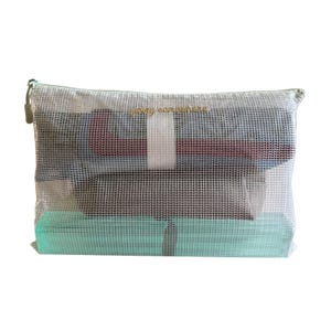 Puede incluir: Un estuche rectangular de malla blanca semitransparente con cremallera y el texto "going somewhere" bordado en color dorado en la parte frontal. El estuche contiene prendas de ropa dobladas en tonos azul oscuro, granate y verde menta.