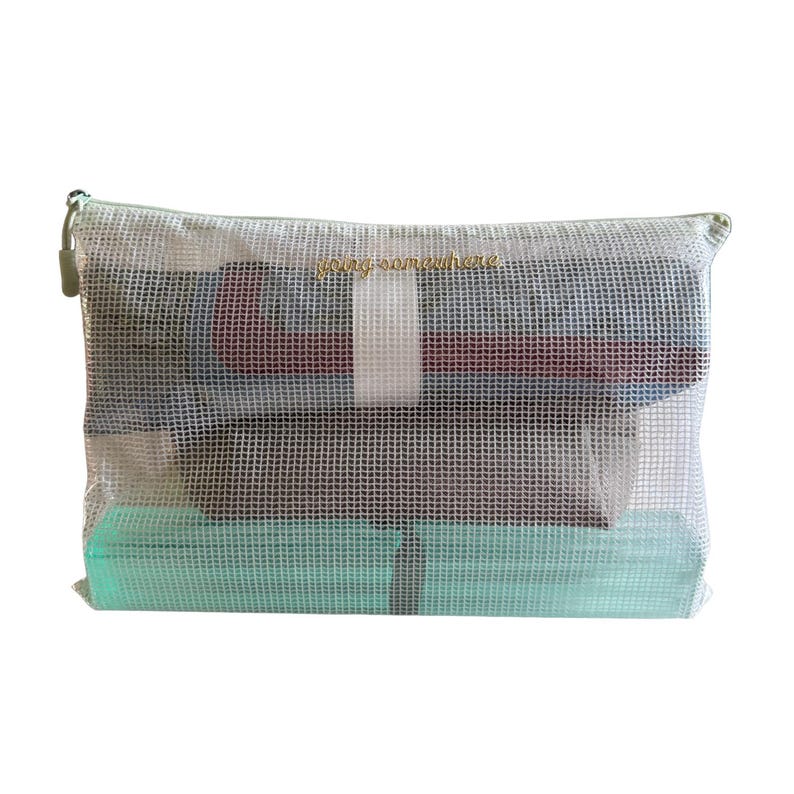 Puede incluir: Un estuche rectangular de malla blanca semitransparente con cremallera y el texto "going somewhere" bordado en color dorado en la parte frontal. El estuche contiene prendas de ropa dobladas en tonos azul oscuro, granate y verde menta.