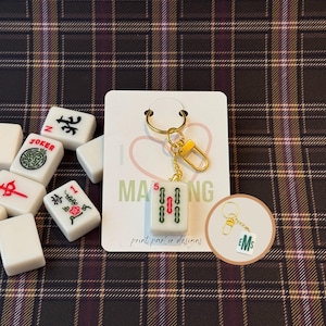 Puede incluir: Un llavero de ficha de Mahjong blanca con un cierre y cadena dorados. La ficha muestra el número 5 y símbolos rojos y verdes. Otras fichas de Mahjong están esparcidas sobre un fondo a cuadros.