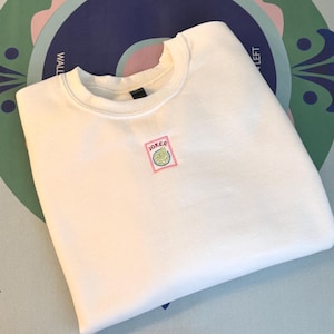 Puede incluir: Una sudadera blanca de cuello redondo con un pequeño parche rectangular bordado en el centro del pecho. El parche tiene un borde rosa y contiene la palabra JOKER en letras rojas sobre un gráfico circular que representa una figura verde y amarilla.