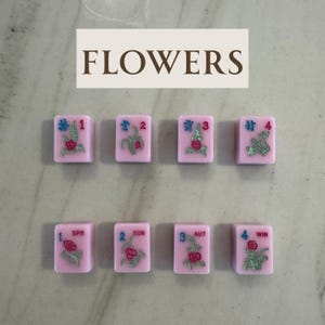 May include: Pink Mahjong tiles with floral designs and numbers 1-4, plus SPR, SUM, AUT, and WIN. The word "FLOWERS" is displayed above the tiles in brown letters. The tiles are arranged in two rows of four.
