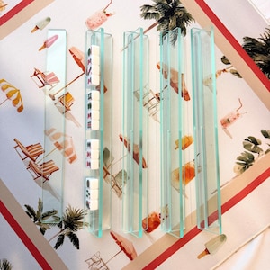 May include: Clear acrylic mahjong tile holders with white tiles, set against a beach-themed patterned background. The background features illustrations of beach chairs, umbrellas, and ice cream. The holders are rectangular and stand upright.