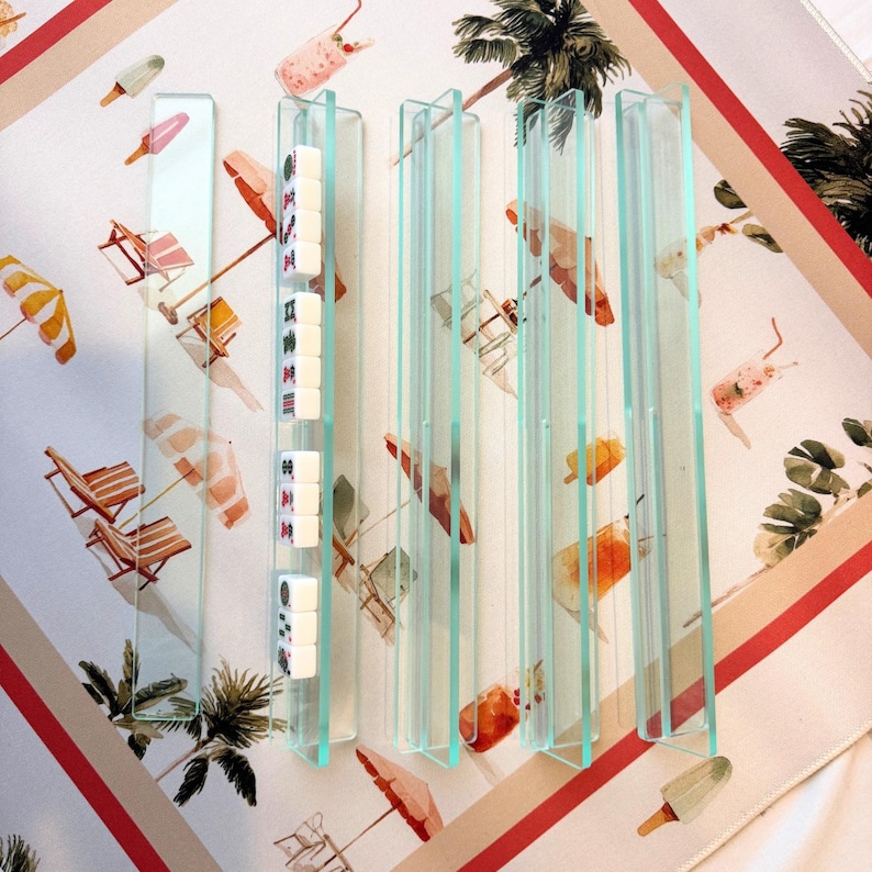 May include: Clear acrylic mahjong tile holders with white tiles, set against a beach-themed patterned background. The background features illustrations of beach chairs, umbrellas, and ice cream. The holders are rectangular and stand upright.