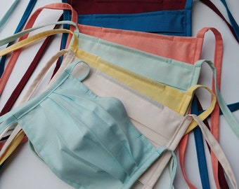 May include: A stack of colorful fabric face masks with different colored ties. The masks are made of cotton and have a pleated design. The masks are arranged in a pile, with the top mask being a light blue color.