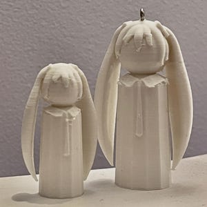 May include: Two white, 3D-printed figurines of a person with long, drooping ears. The figurines have a simple, geometric design with a cylindrical body and a head with a small, rounded crown. One figurine is smaller than the other.
