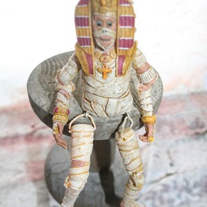 May include: A mummy figurine wearing a yellow and red headdress with a gold ankh pendant. The figurine is sitting on a wooden base.